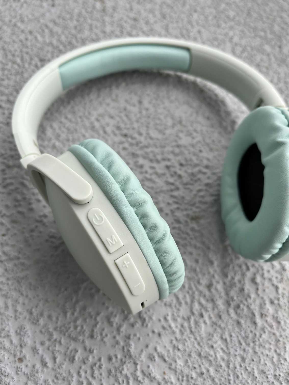 Close-up of mint green Bluetooth headphones showing control buttons and soft ear cushions, displayed on a textured gray background.