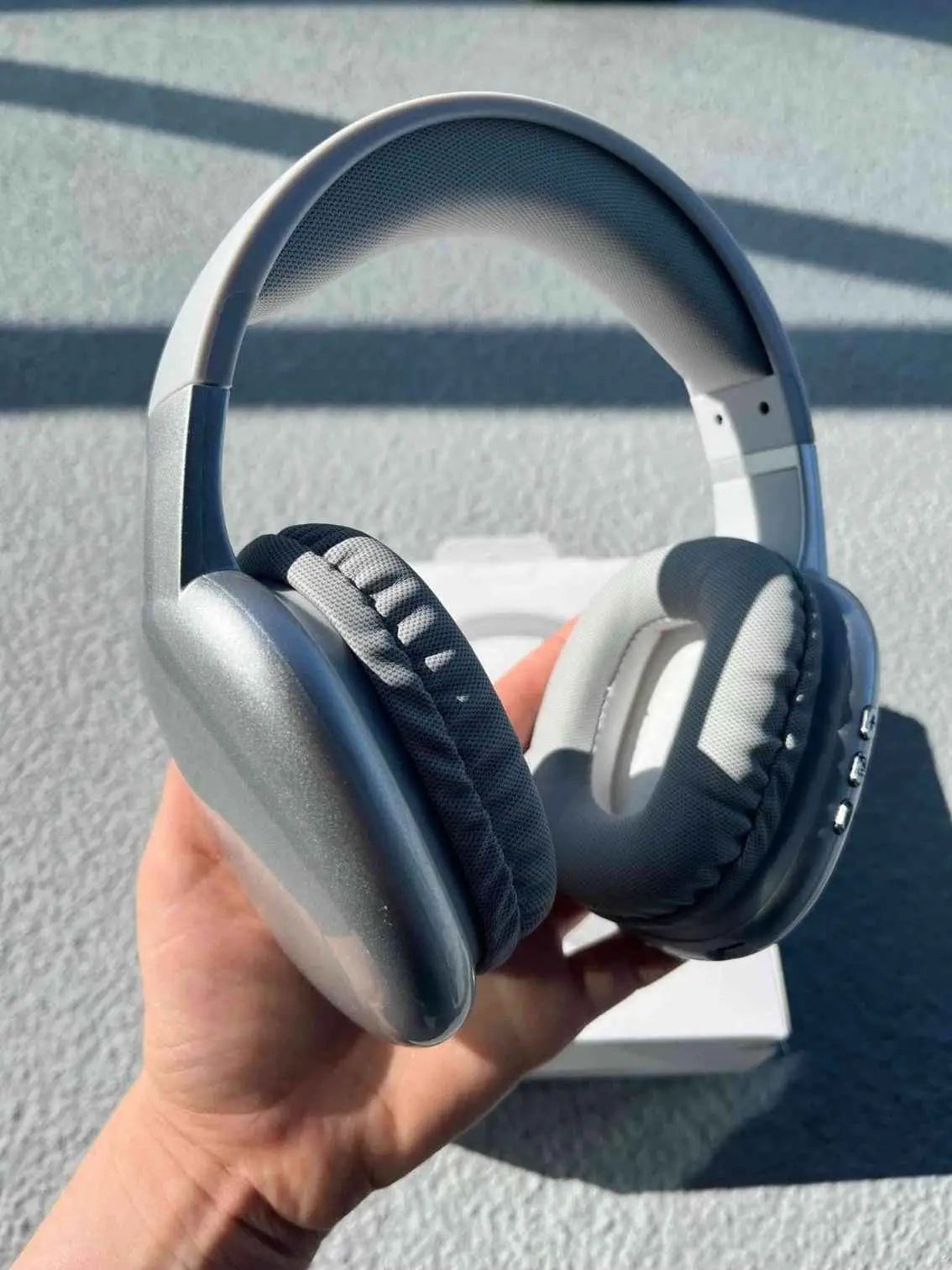 Silver PG02 over-ear Bluetooth headphones with cushioned ear pads, held in hand with packaging in the background.